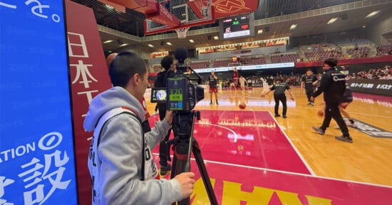 A cameraman films a basketball game from the sidelines inside an indoor arena, where players are warming up on the court and spectators are seated in the stands.