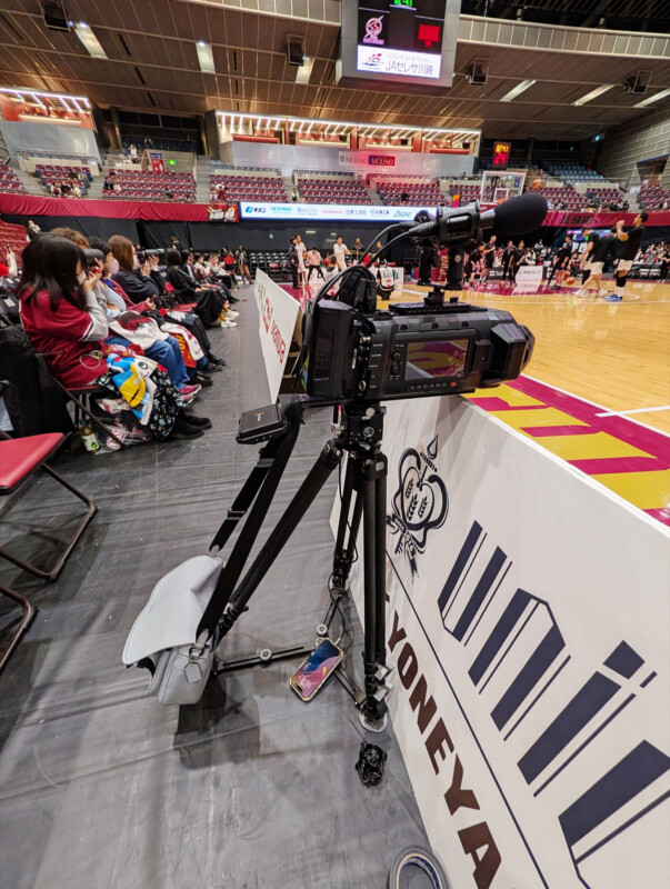 A professional video camera is set up courtside on a tripod, pointing toward a basketball court during a game. Spectators sit in the background alongside the court, with arena seats visible further back.