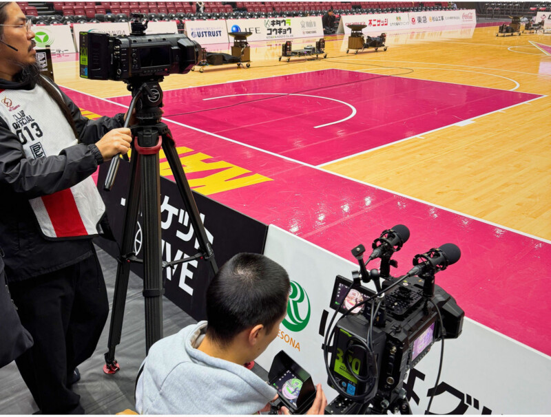 Two camera operators film an empty indoor basketball court from the sidelines, focusing their cameras on the court. The arena seats in the background are unoccupied.