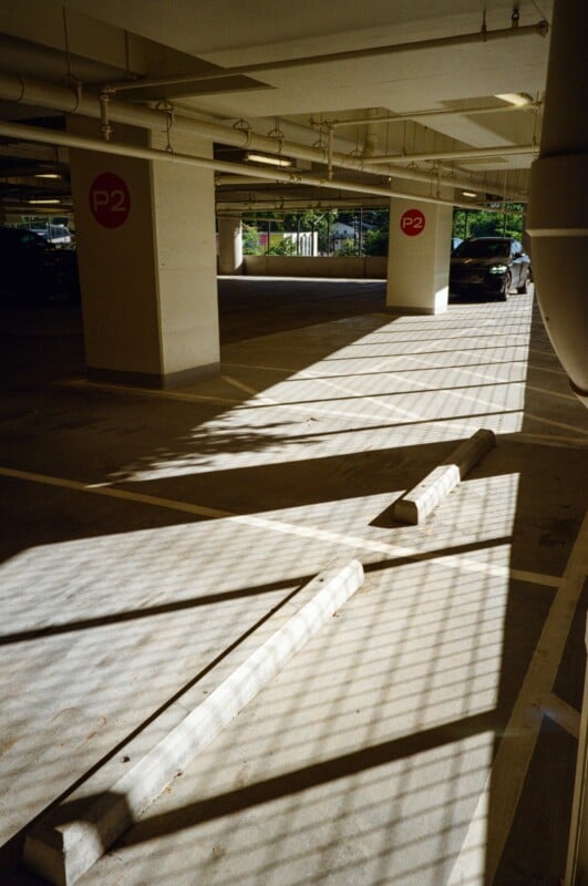 Sunlight streams through a fence, casting striped shadows on the floor of a mostly empty parking garage. White concrete pillars marked "P2" and a parked black car are visible in the background.
