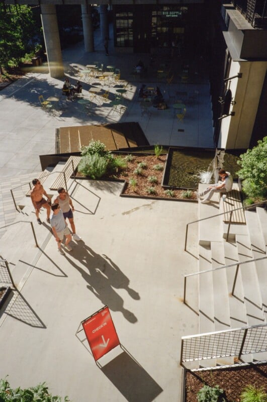 A sunny outdoor patio with people relaxing on chairs and walking. Three people walk past a red sign that says "CHECK-IN" with an arrow. Another person sits on steps near a small fountain and plants.