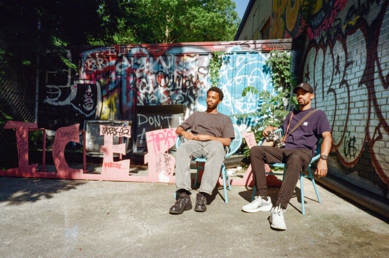 Two people sit on chairs in front of a wall covered in colorful graffiti. They are outdoors on a sunny day, with shadows on the ground and trees in the background. Both appear relaxed, looking toward the camera.