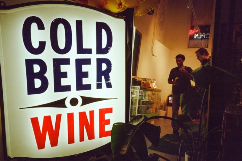 Large illuminated sign reading "COLD BEER WINE" in a window at night; two people are standing and talking inside the warmly lit shop with a neon "OPEN" sign in the background.