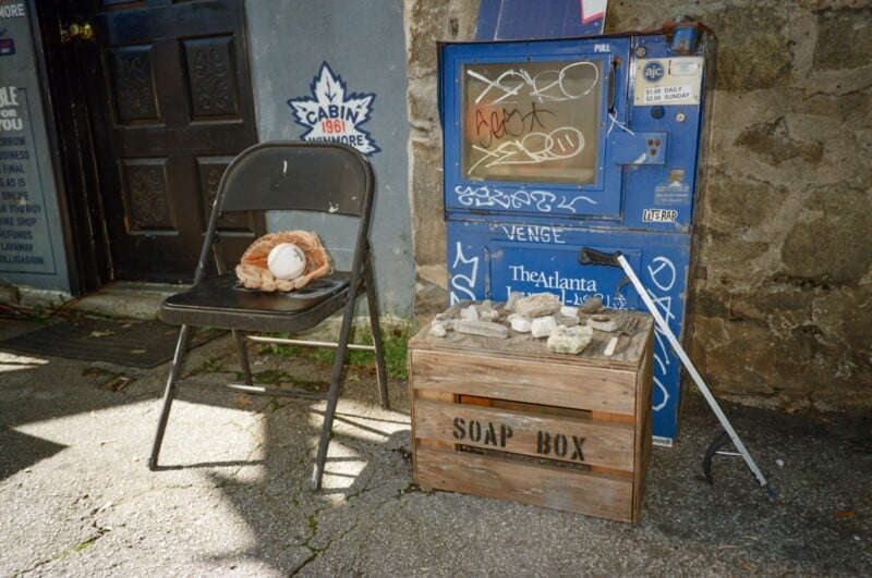 A black folding chair with a baseball glove and ball sits beside a blue, graffiti-covered vending machine. A wooden crate labeled "SOAP BOX" holds rocks, and a grabber tool leans nearby in a sunlit alley.