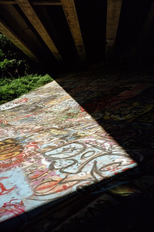 A beam of sunlight illuminates colorful graffiti on a concrete surface beneath a bridge, with shadows from the bridge overhead and greenery visible at the edge.