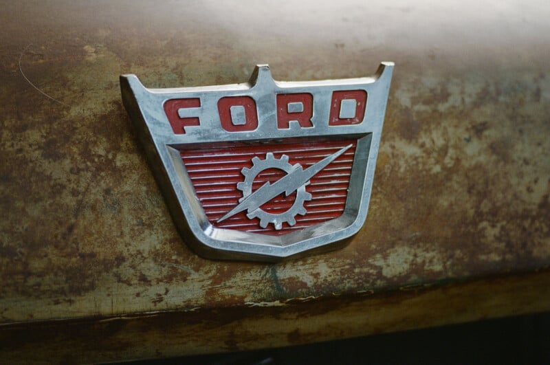 Close-up of a vintage Ford emblem featuring a red and silver badge with a gear and lightning bolt design, mounted on a rusted, weathered car surface.