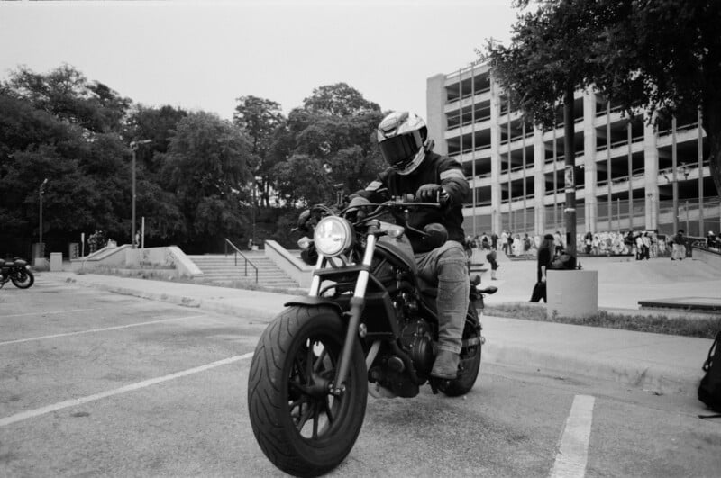 A person wearing a helmet and protective gear rides a motorcycle in a parking lot, with a multi-level parking garage and trees in the background.
