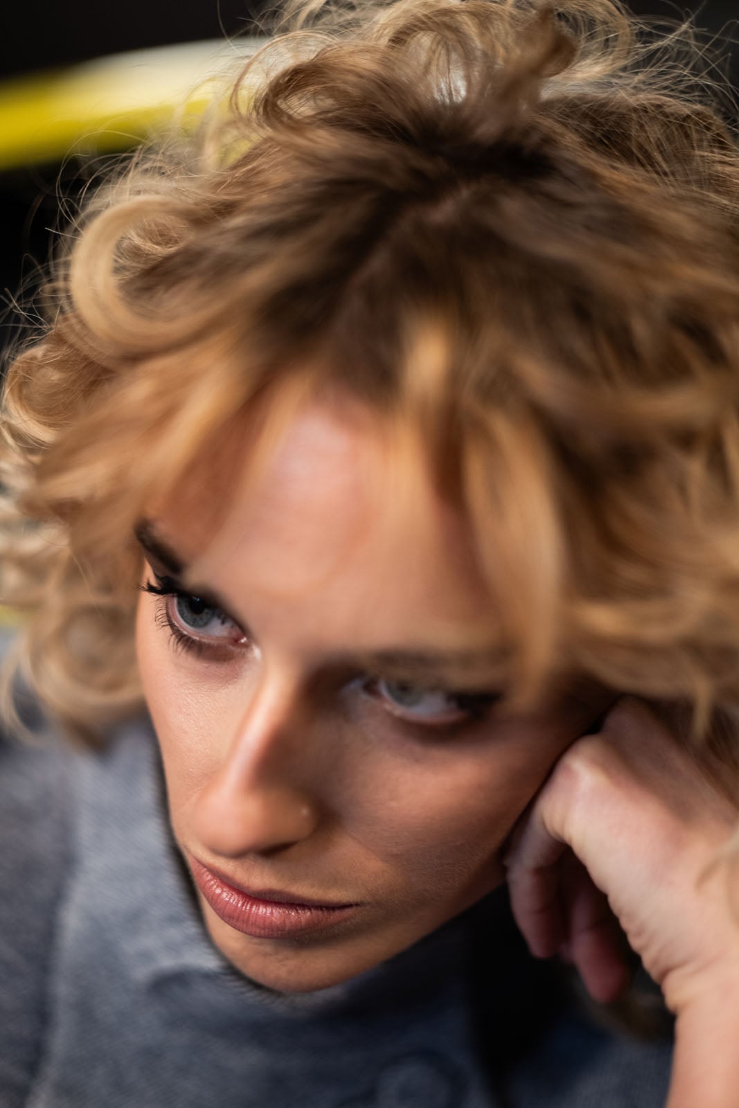 A person with curly blonde hair and blue eyes looks down thoughtfully, resting their chin on their hand. The image is softly focused and closely cropped, highlighting their facial expression.