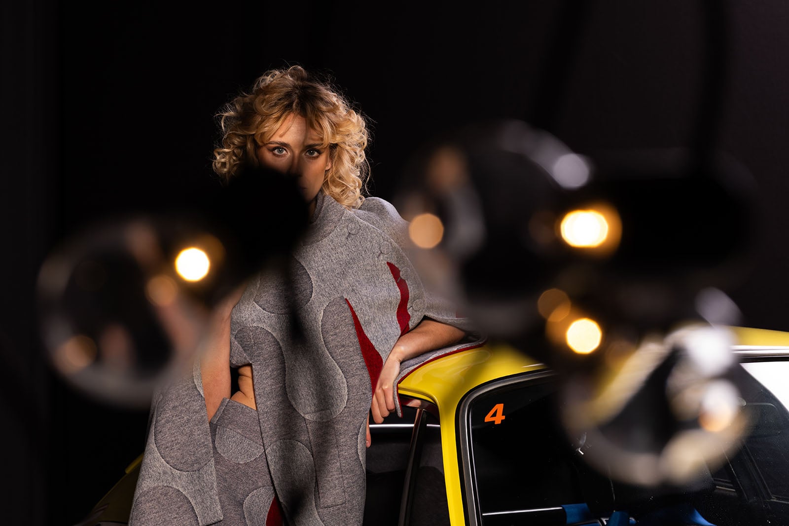 A woman with curly blonde hair stands next to a yellow car, partially obscured by out-of-focus hanging lights in the foreground. She is wearing a gray patterned garment and gazes directly at the camera.