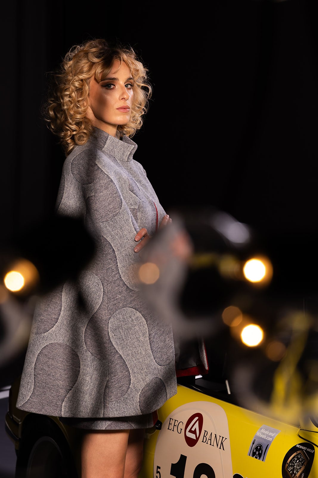 A woman with curly blonde hair wears a textured grey coat and stands confidently beside a yellow car with EFG Bank logos, with blurred lights in the foreground against a dark background.