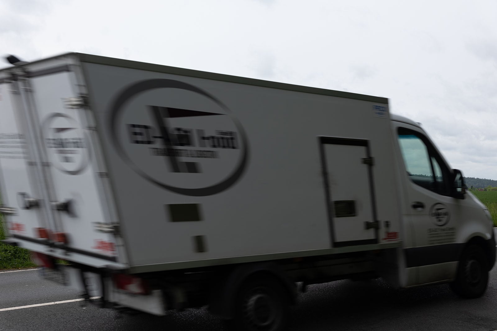 A blurred delivery truck with a large circular logo on its side is driving on a road. The surrounding environment appears overcast and slightly rural. The text on the truck is not clearly visible.