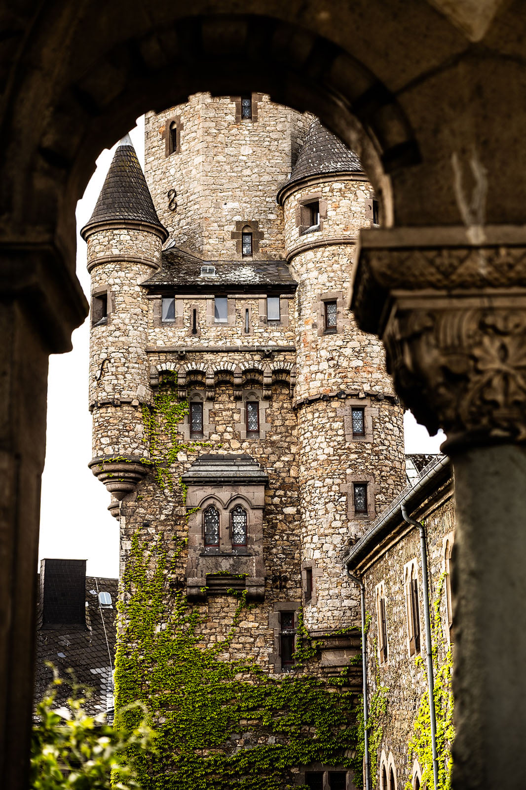A stone castle tower with ivy climbing its walls is framed by an ornate archway in the foreground. The tower features arched windows, turrets, and a steep slate roof.