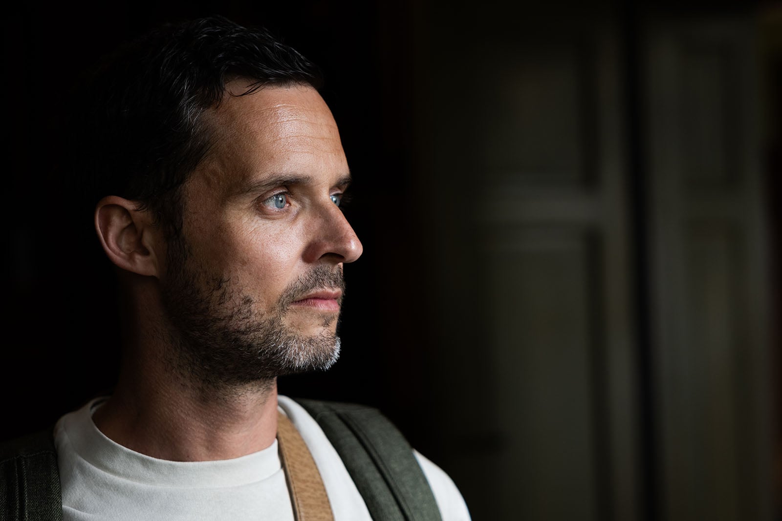 A man with short dark hair and a beard gazes thoughtfully to the side in soft lighting, wearing a white shirt and backpack, with a blurred background featuring light-colored doors.