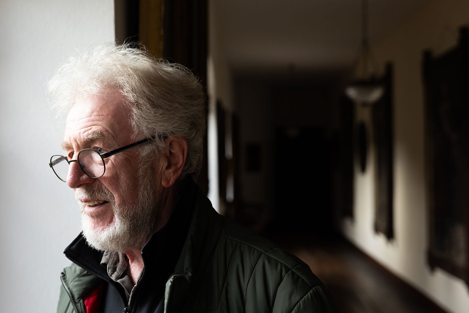 An older man with white hair, a beard, and glasses stands by a window, smiling softly. He wears a dark jacket and looks outside, with a dimly lit hallway stretching out behind him.