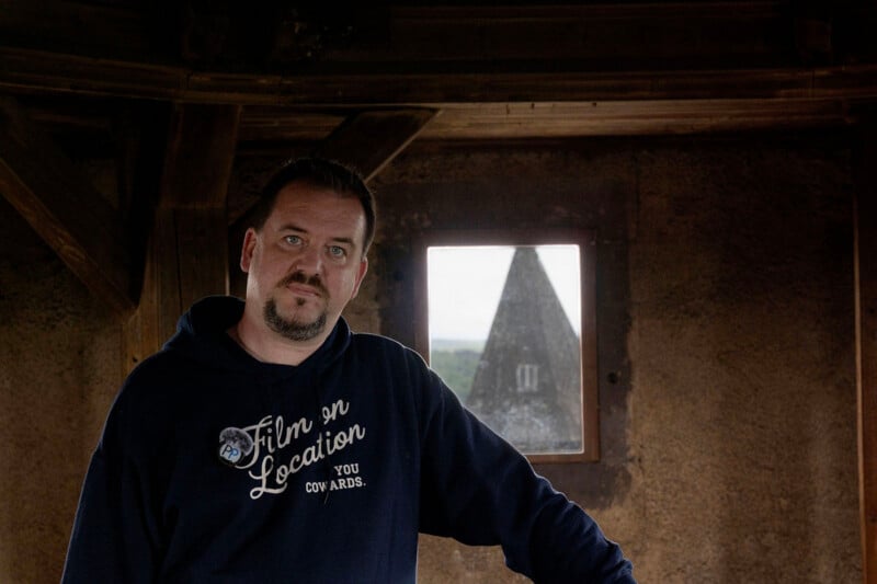 A man with short hair and a beard, wearing a navy blue "Film on Location" hoodie, stands inside a dimly lit rustic room with wooden beams. A small window behind him shows a blurry, pointed-roof building outside.