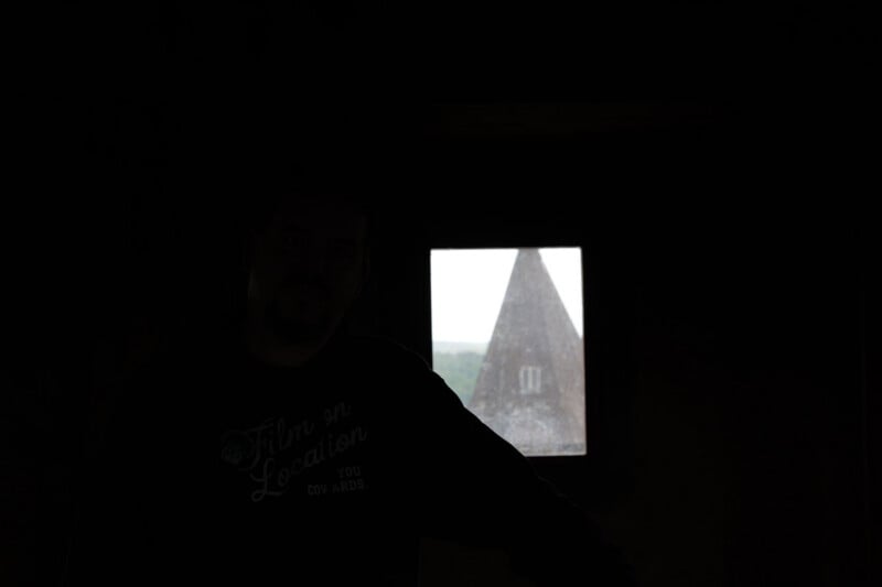 A person stands in a dark room with a small window behind them, through which a pointed tower and part of a landscape are visible in daylight. The person's features are mostly obscured by shadow.