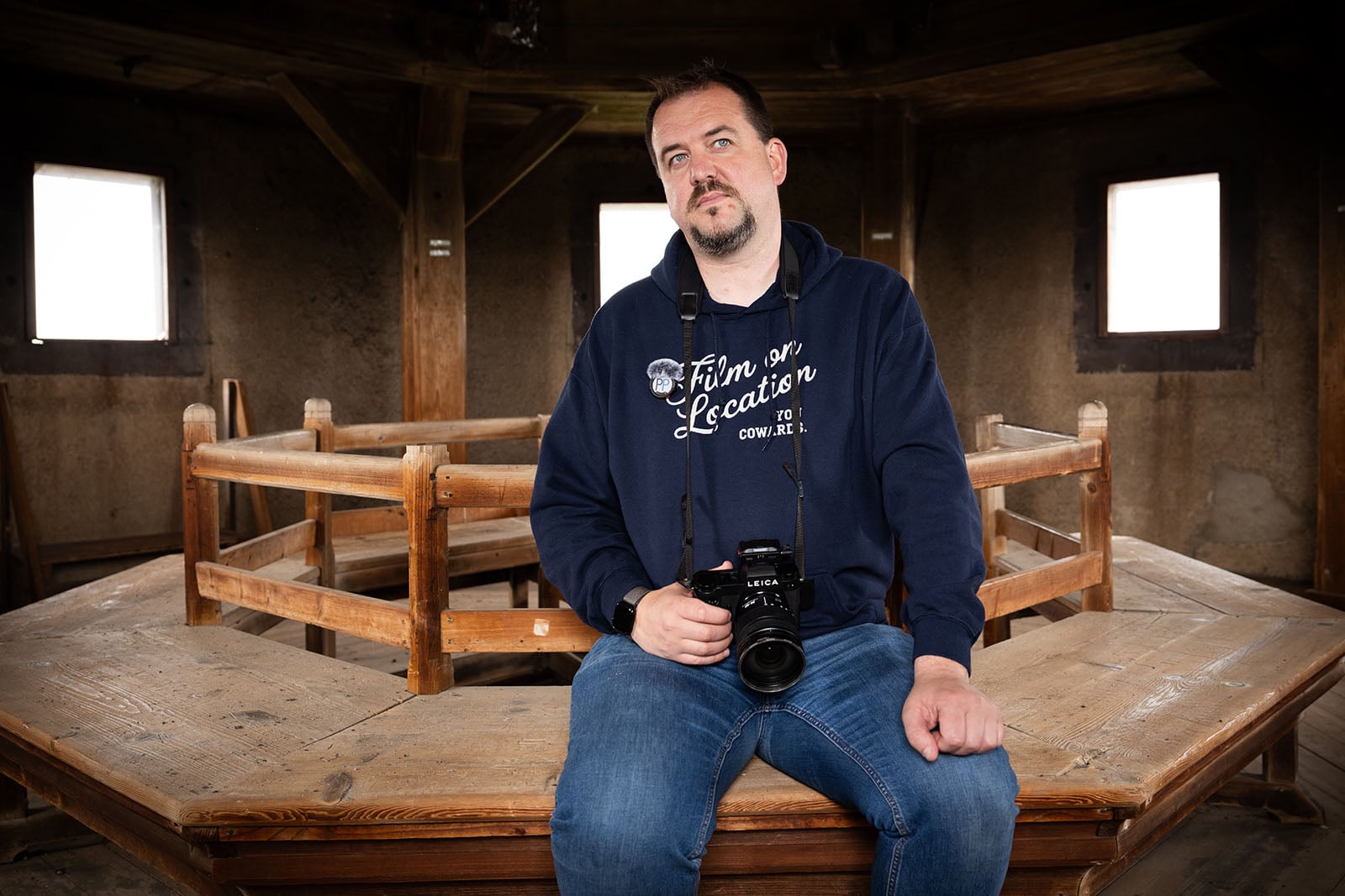 A man in a navy hoodie and jeans sits on a wooden structure indoors, holding a camera. He looks slightly upward with a thoughtful expression. The room has wooden beams and windows with white light outside.
