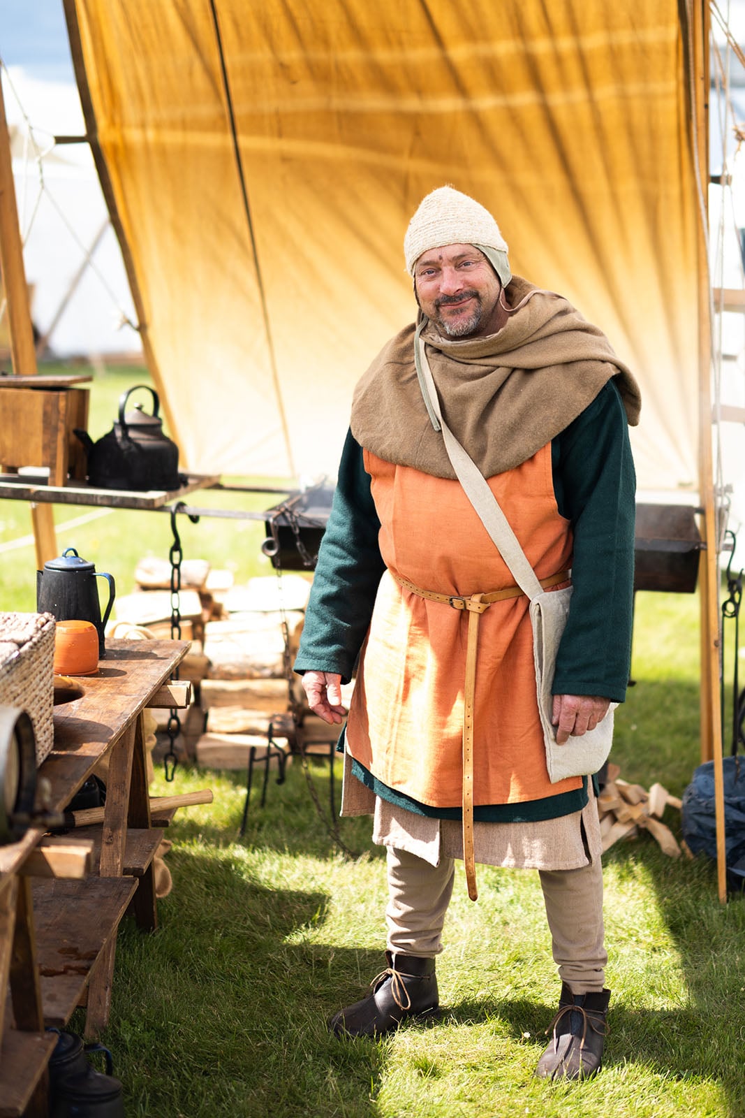 A man in medieval costume stands smiling under a yellow tent at an outdoor reenactment camp, surrounded by wooden tables, cooking pots, and other historical props on a grassy field.