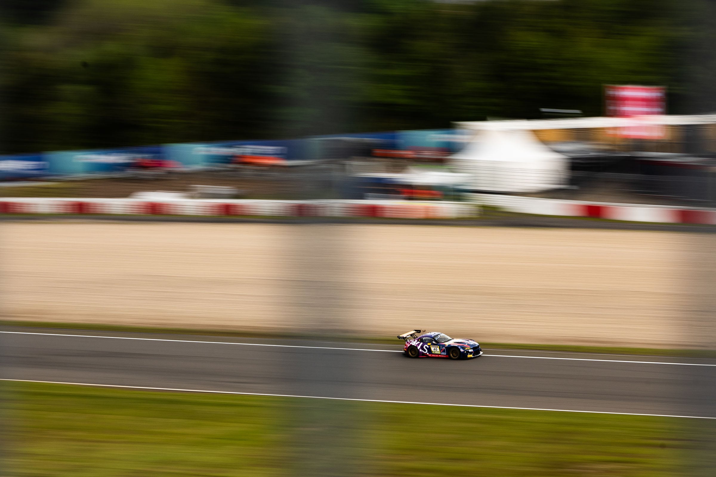 A race car speeds along a track, with motion blur emphasizing its rapid movement. The background shows barriers, blurred spectators, and tents, with green trees beyond. Vertical bars partially obscure the view.
