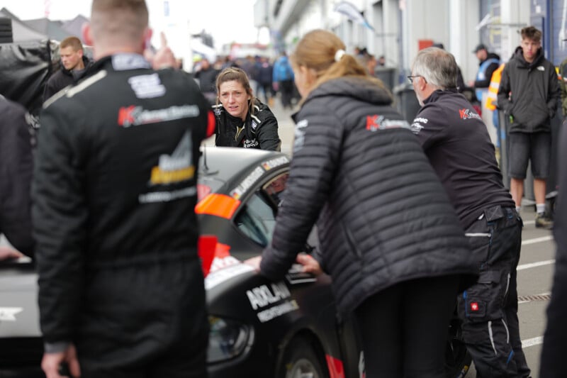 A race car crew in black jackets and gear works around a black car in the pit area, while a woman in the center looks intently at the vehicle. Other people and cars are visible in the busy background.