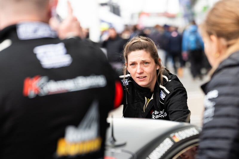 A woman in a racing suit stands by a car, looking intently at someone off-camera. Two other people in racing gear are nearby, and a blurred crowd is visible in the background at what appears to be a racing event.