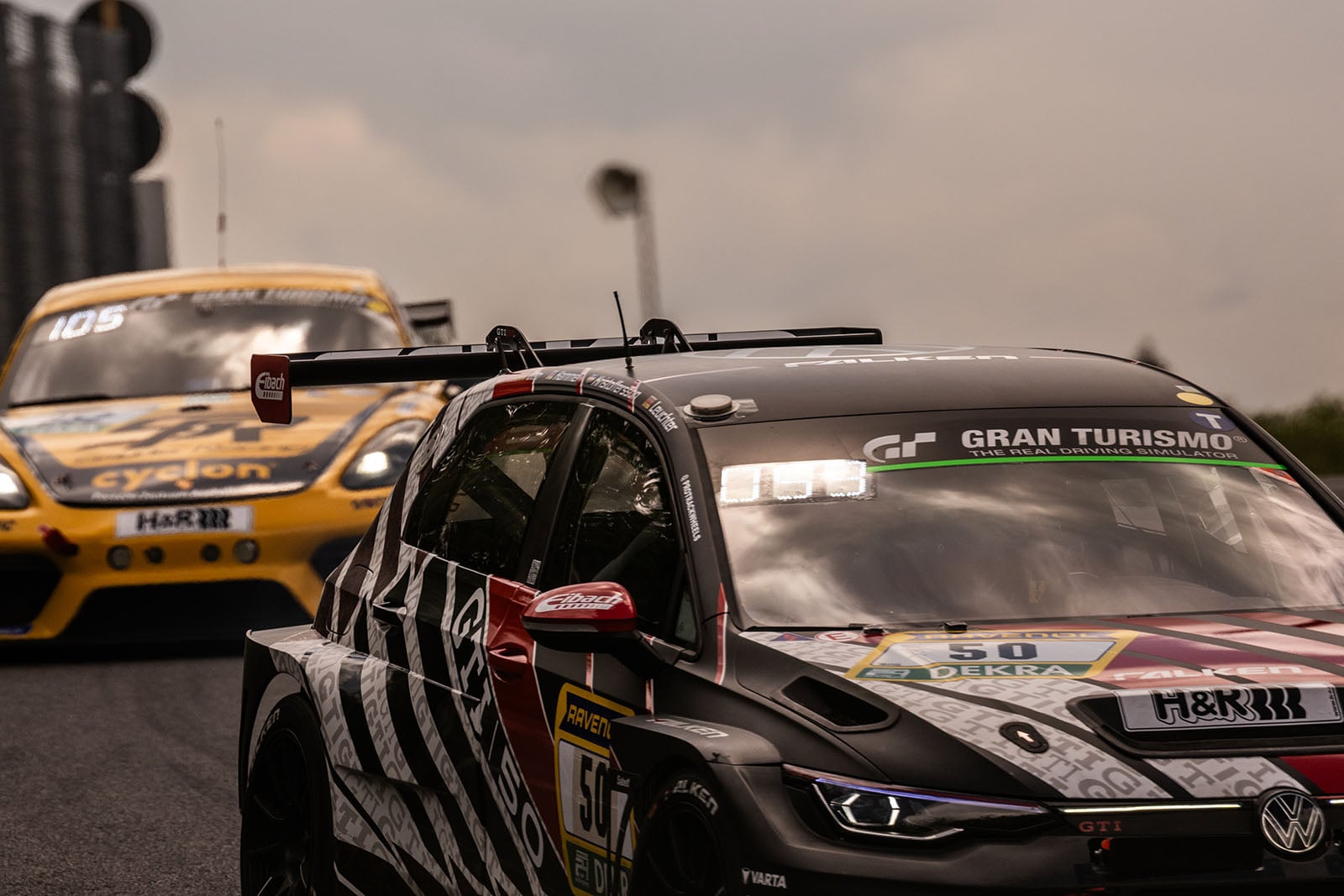 A close-up of two racing cars on a track, with a black and red car in the foreground and a yellow car following close behind under a cloudy sky. Both cars have visible sponsor logos and racing decals.