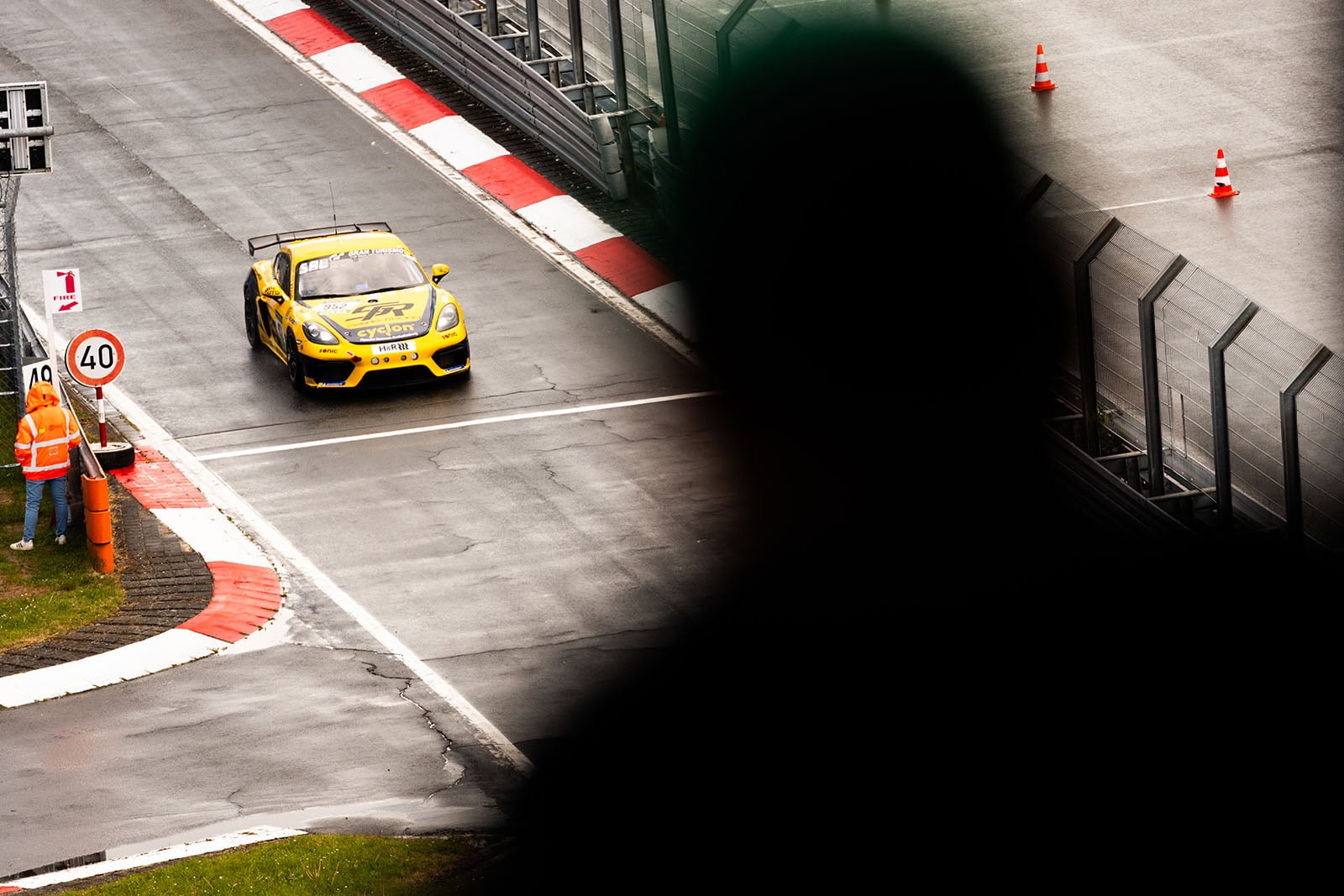 A yellow race car drives on a wet racetrack near a curve, as a person’s silhouette partially obscures the view. Safety cones and a 40 km/h sign are visible; an official in an orange vest stands by the track.