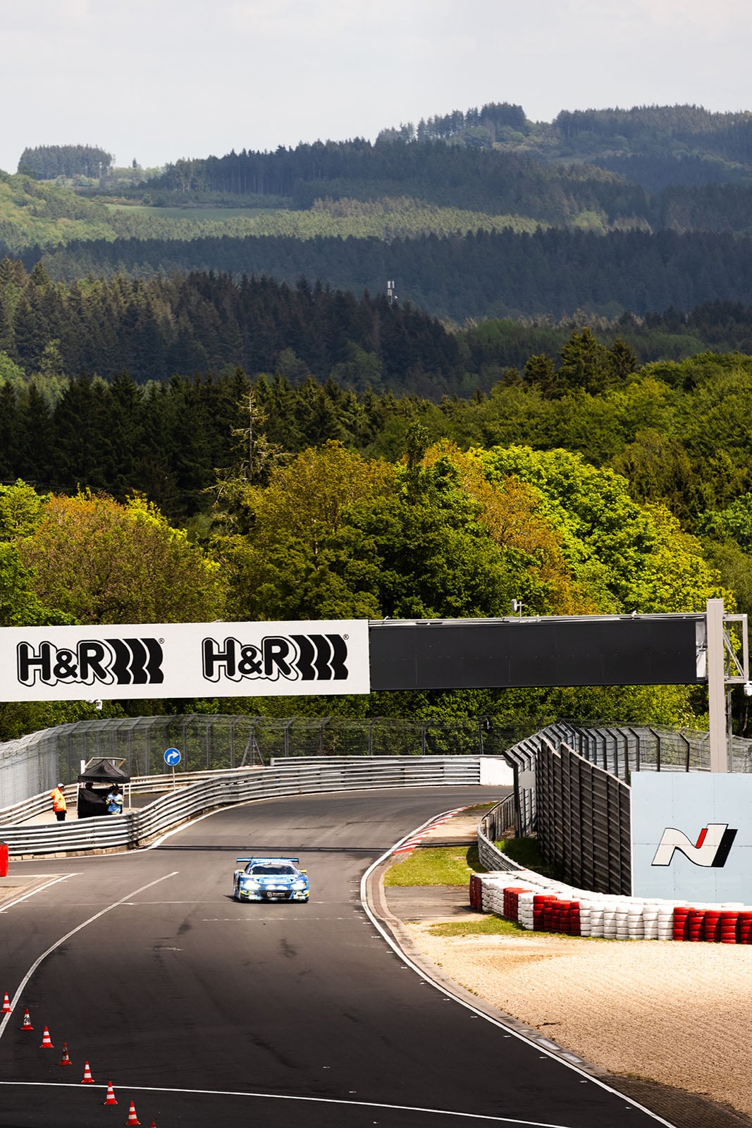 A blue race car drives on a winding race track surrounded by green trees and hills, passing under a bridge with "H&R" signs on a sunny day.