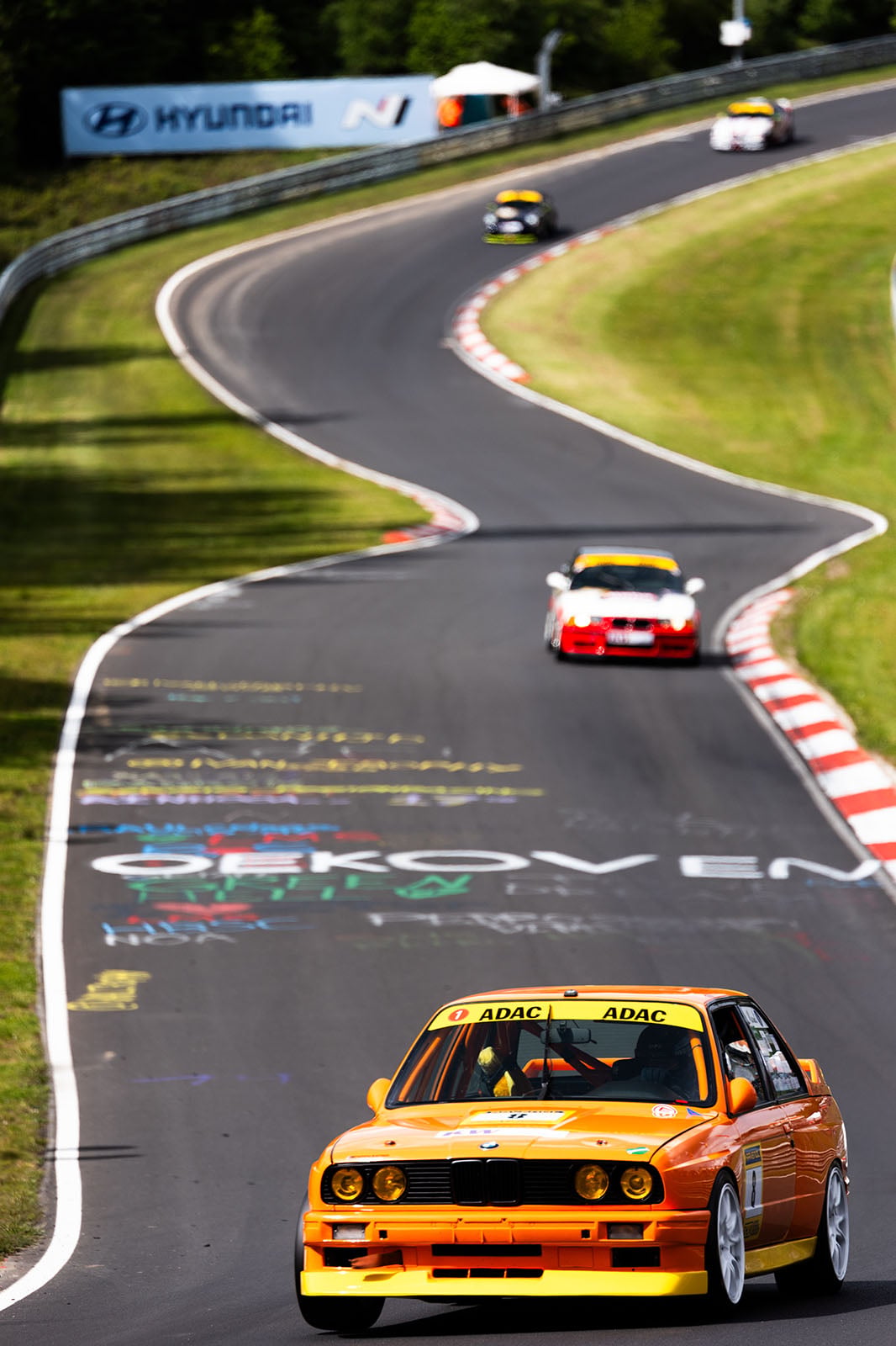 A yellow race car leads several other cars around a winding racetrack, surrounded by greenery and colorful graffiti on the track surface. A Hyundai banner is visible in the background.