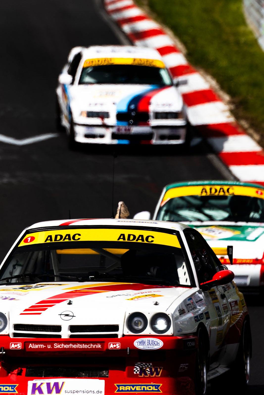 Three race cars with ADAC banners drive on a curving racetrack, bordered by red and white curbs and green grass. The focus is on the leading white car, with two more cars close behind.