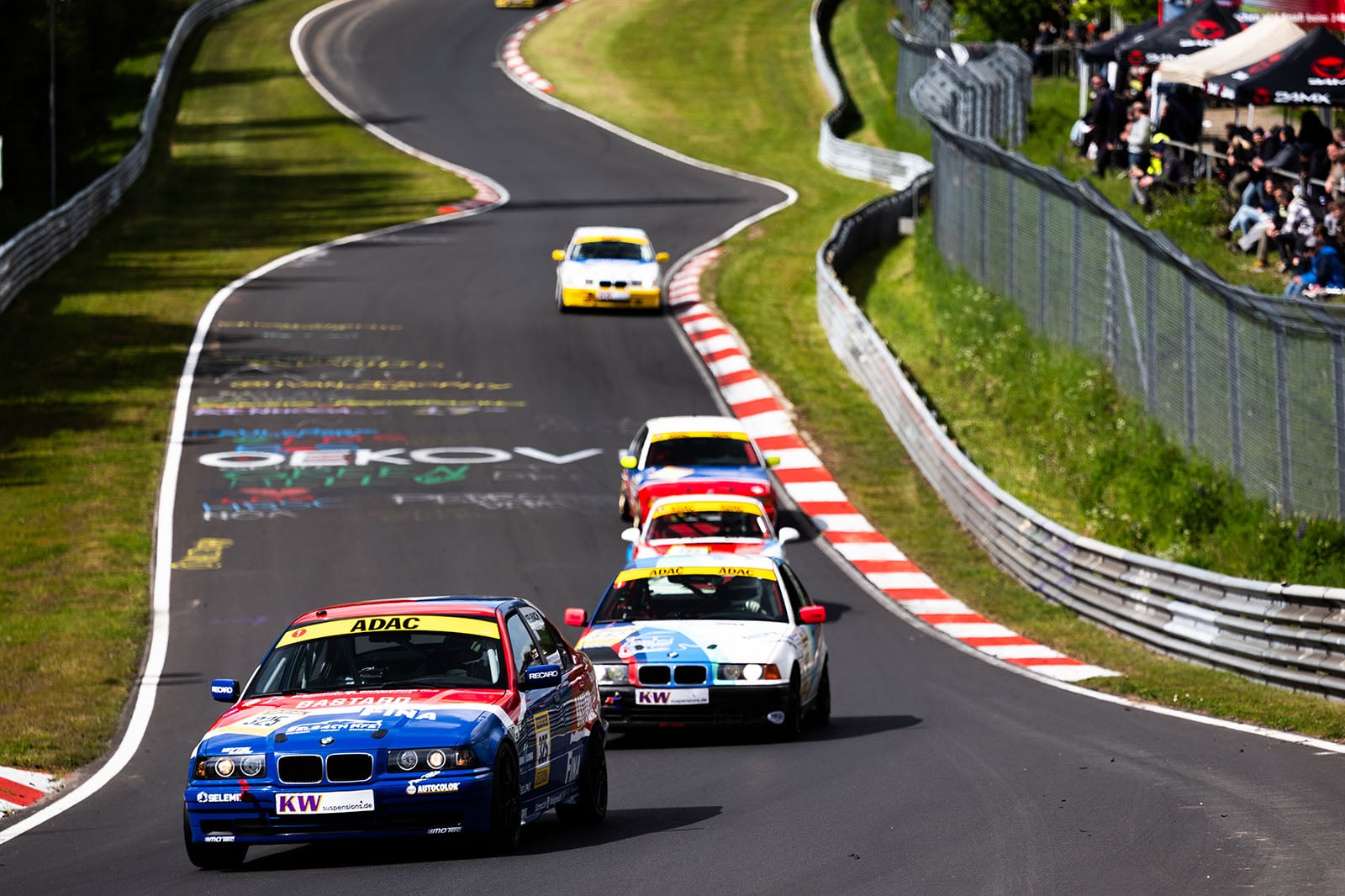 Four race cars speed along a winding track lined with grass and safety barriers, with spectators watching from the right. The leading car is blue, followed by white and yellow cars under sunny skies.
