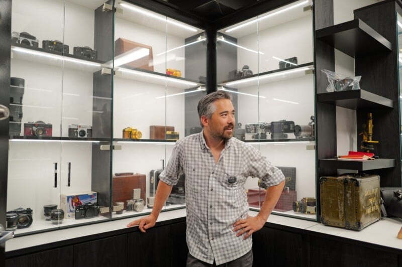 A man with gray hair and a beard stands in front of glass display cases filled with vintage cameras, camera equipment, and collectibles in a well-lit room.