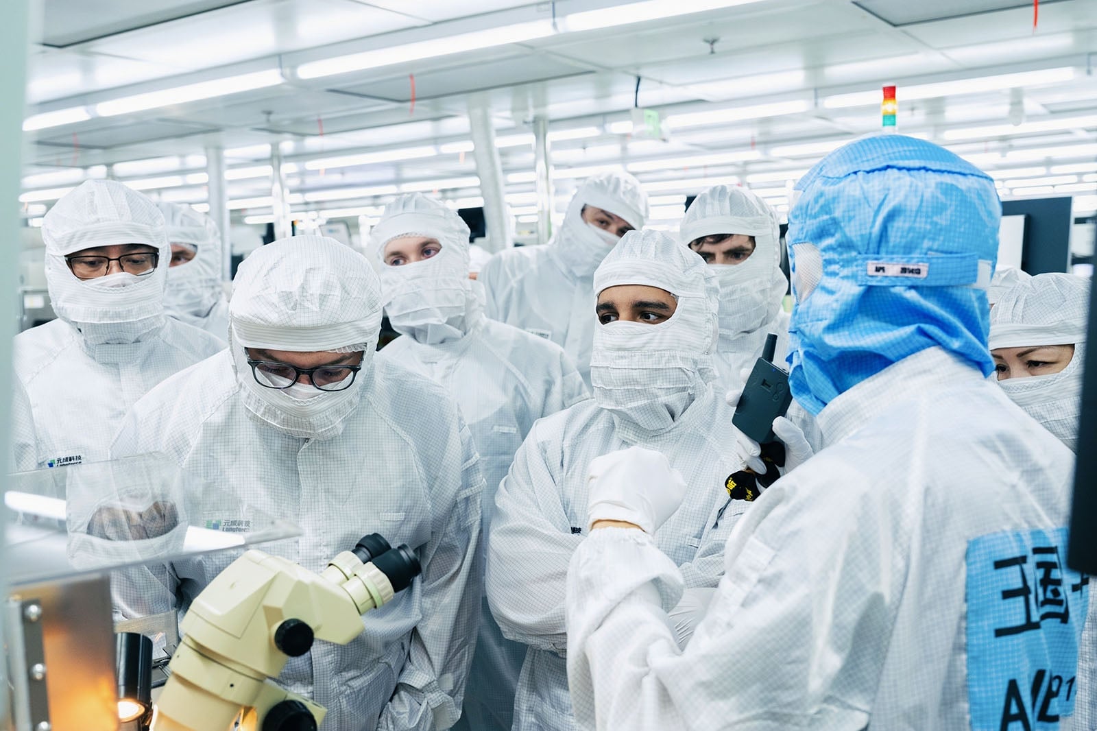 A group of people in white cleanroom suits and face masks listen to a person in a blue suit explaining something near lab equipment and a microscope in a high-tech laboratory setting.