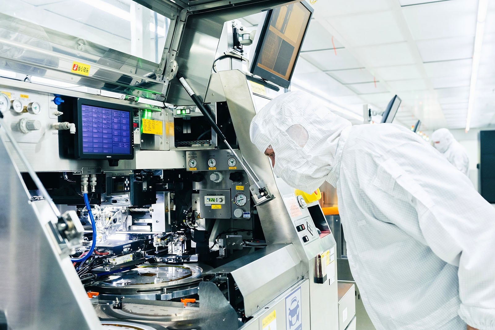 A person in a cleanroom suit closely inspecting high-tech machinery in a laboratory or semiconductor manufacturing environment. Monitors and equipment surround the individual in the sterile, brightly lit room.