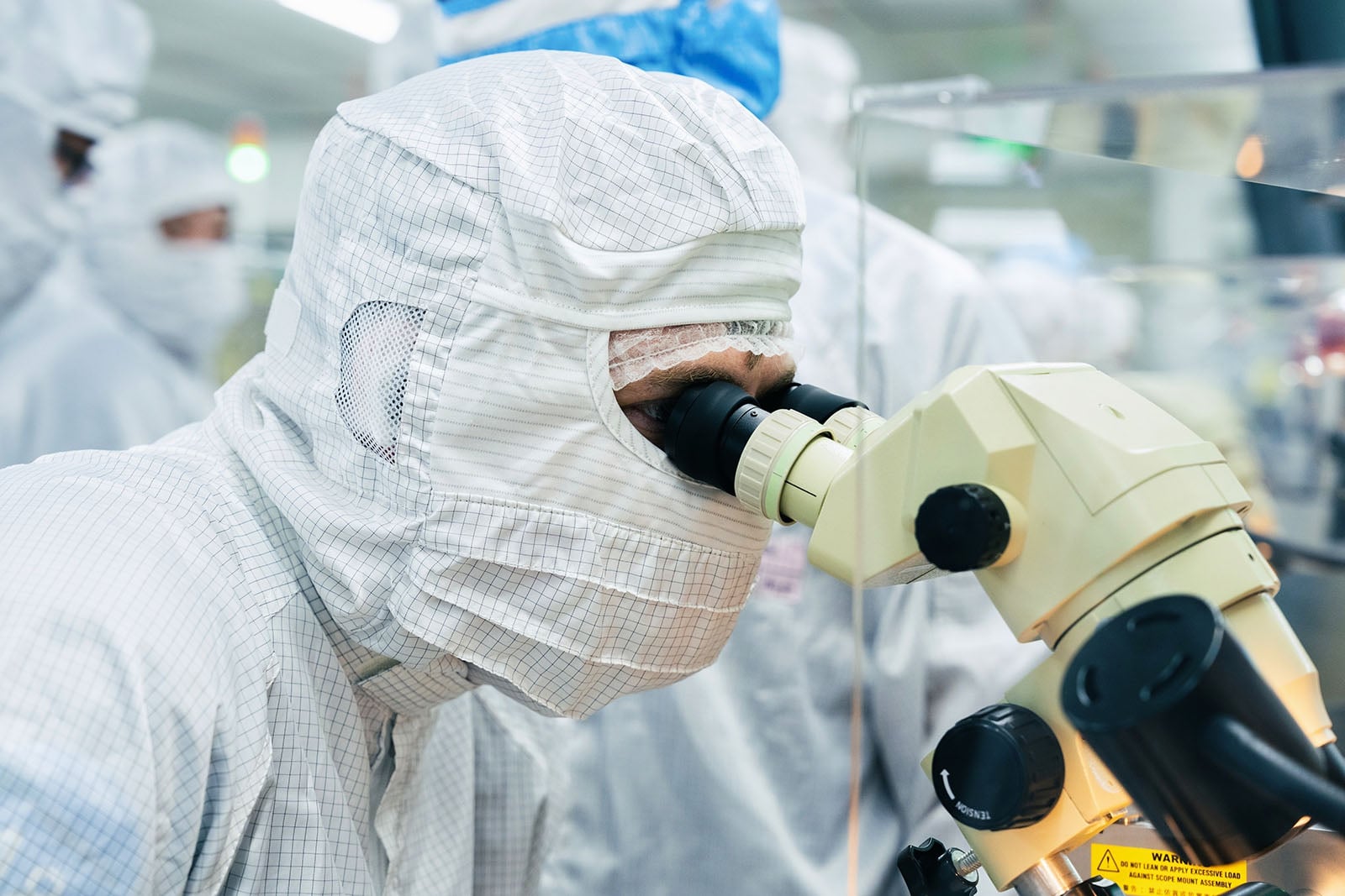 A scientist wearing a full-body cleanroom suit and mask looks into a microscope in a laboratory environment, with other similarly dressed individuals working in the background.