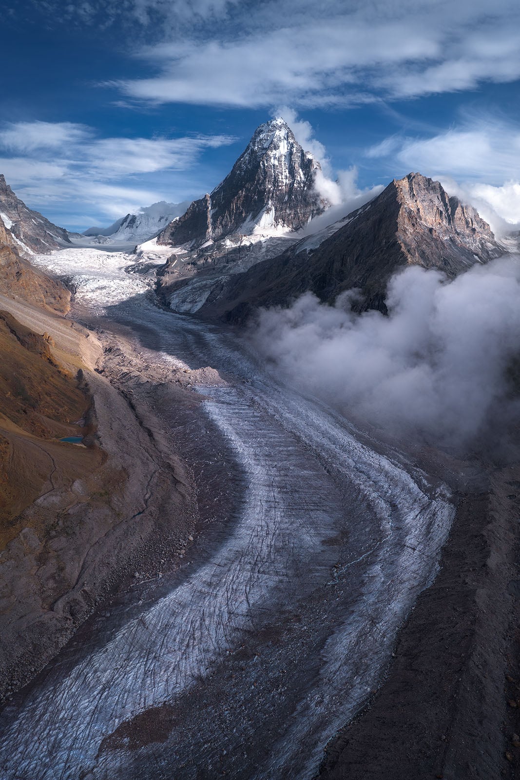 A winding glacier flows between rugged, rocky mountains under a blue sky with scattered clouds. Snow caps the highest peak in the background, while mist rises from the valley below.
