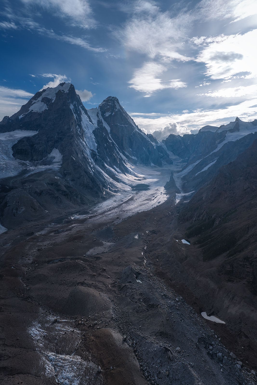 A sweeping mountain landscape with rugged, rocky peaks capped by snow and glaciers. A winding, rocky valley stretches toward the foreground under a partly cloudy blue sky.