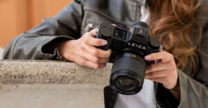 A person wearing a brown leather jacket holds a Leica camera with both hands, aiming it over a stone ledge. The camera’s buttons, dials, and lens are clearly visible.