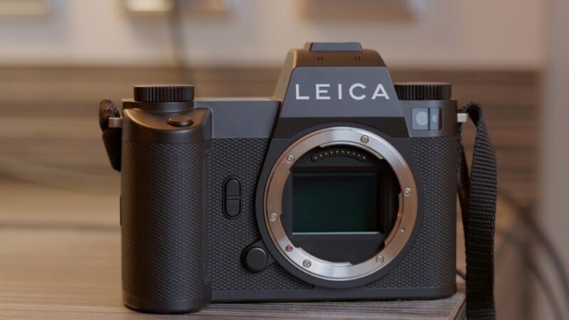 A Leica digital camera without a lens is placed on a wooden surface, showing its front view and sensor. The body is black with textured grips and a visible camera strap on the right.