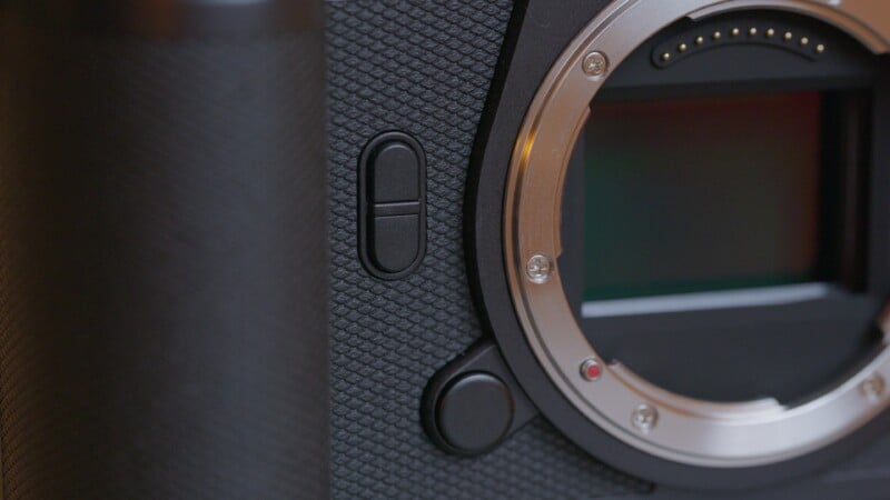Close-up of a digital camera body showing the textured grip, two buttons, and the metal lens mount with visible electronic contacts and part of the camera sensor.