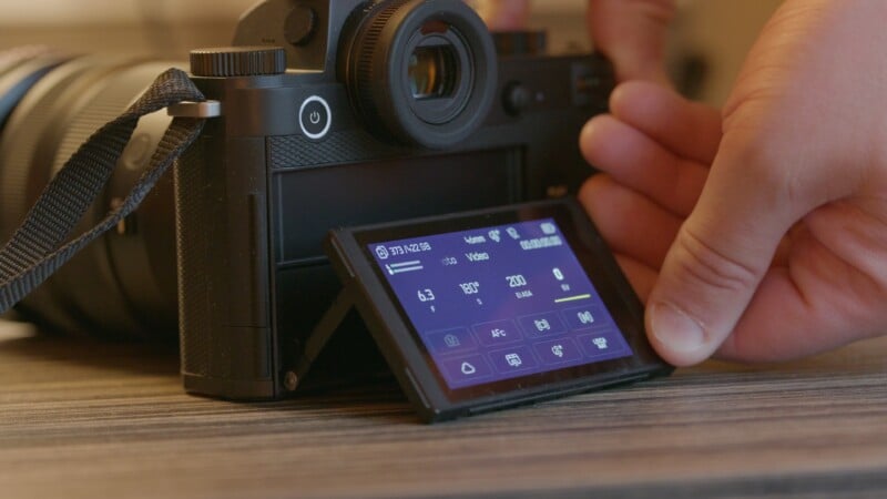 A close-up of a digital camera on a table, with a person tilting the touchscreen display to adjust settings visible on the screen.