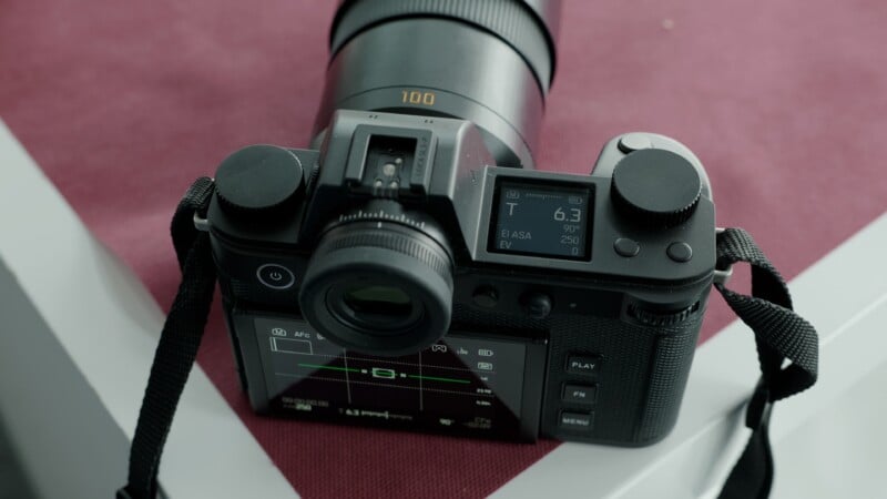 A digital camera with a large telephoto lens sits on a maroon surface; its top display shows settings such as aperture, ISO, and exposure compensation. The rear screen displays additional camera information.