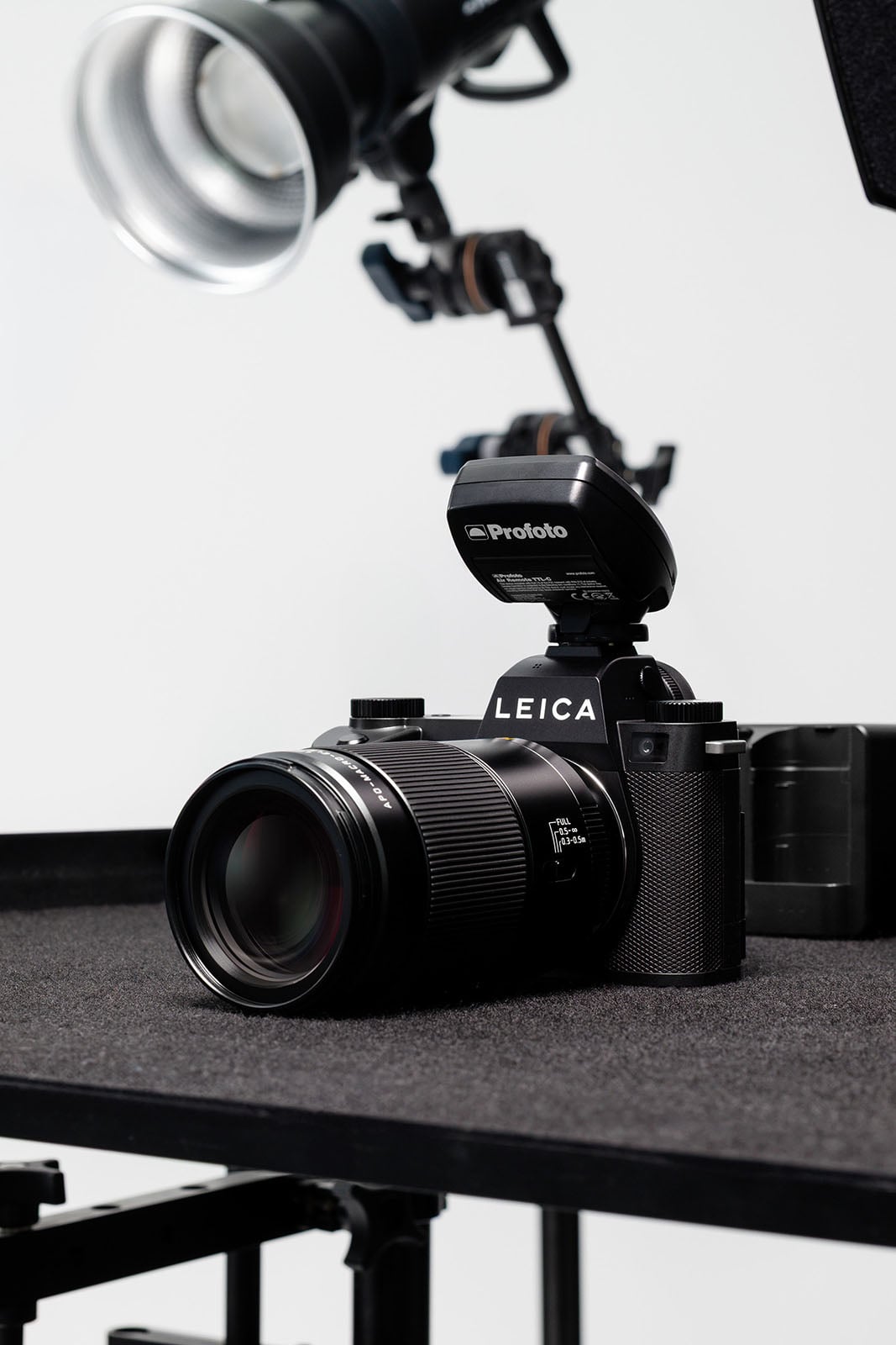 A Leica camera with a large lens and a Profoto flash attached is placed on a black table, with studio lighting equipment visible in the background.