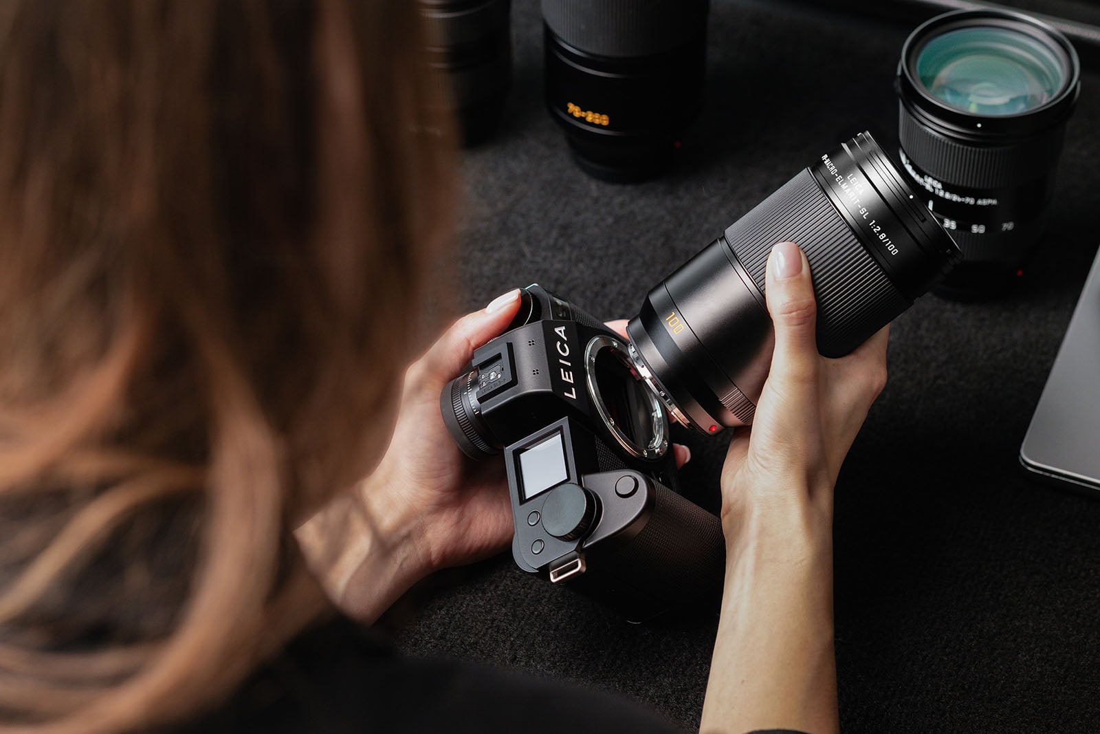 A person holds a Leica camera in one hand and attaches a large lens with the other, surrounded by additional camera lenses on a dark surface.