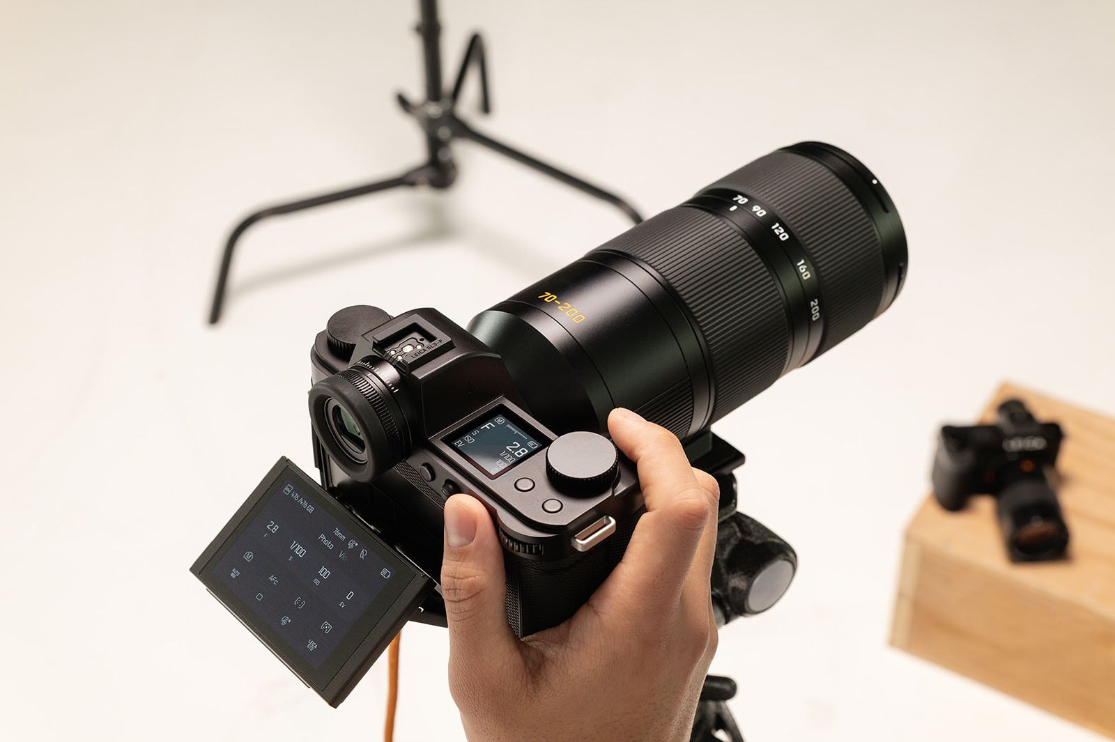 A person holds a digital camera with a large telephoto lens and a flipped-out LCD screen, mounted on a tripod. Another camera rests on a wooden box in the blurred background.