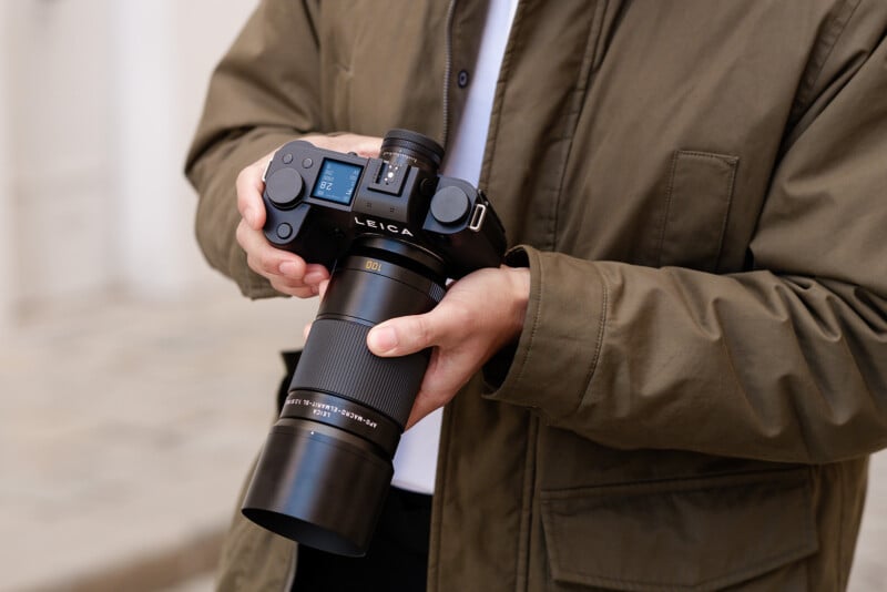 A person in a green jacket holds a Leica camera with a large telephoto lens, adjusting settings on the camera. The background is softly blurred.