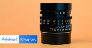 A camera lens with yellow and white distance and aperture markings sits upright on an orange background. The "PetaPixel Reviews" logo appears in the lower left corner.