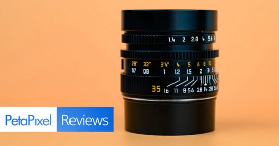 A camera lens with yellow and white distance and aperture markings sits upright on an orange background. The "PetaPixel Reviews" logo appears in the lower left corner.