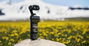 A handheld camera with a screen and dual lenses stands on a rock in a field of yellow flowers, with snow-covered mountains blurred in the background.