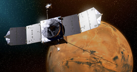 A spacecraft with large solar panels orbits Mars, with the planet’s orange, cratered surface and thin atmosphere visible in the background against a starry space.